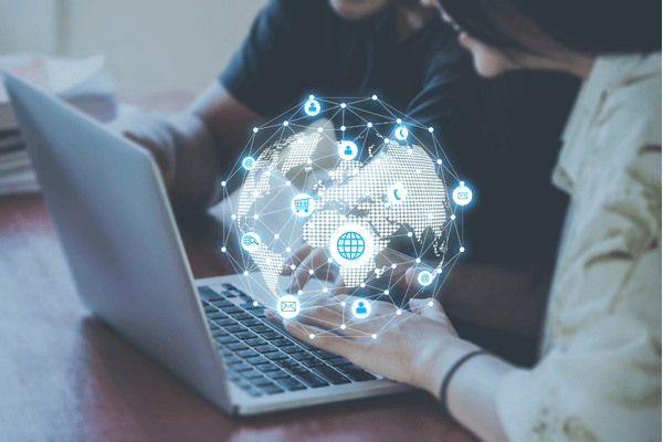 A digital globe with interconnected icons representing global communication and commerce hovers over a laptop keyboard, with hands interacting with the interface.